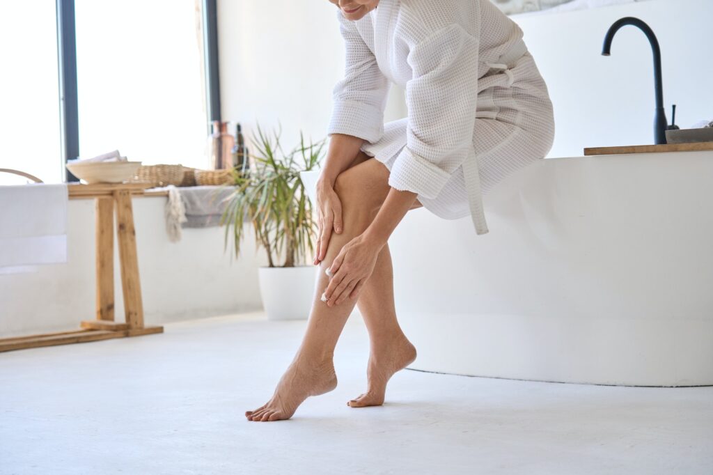 Middle aged mature woman applying veins crème on legs sitting in bathroom.